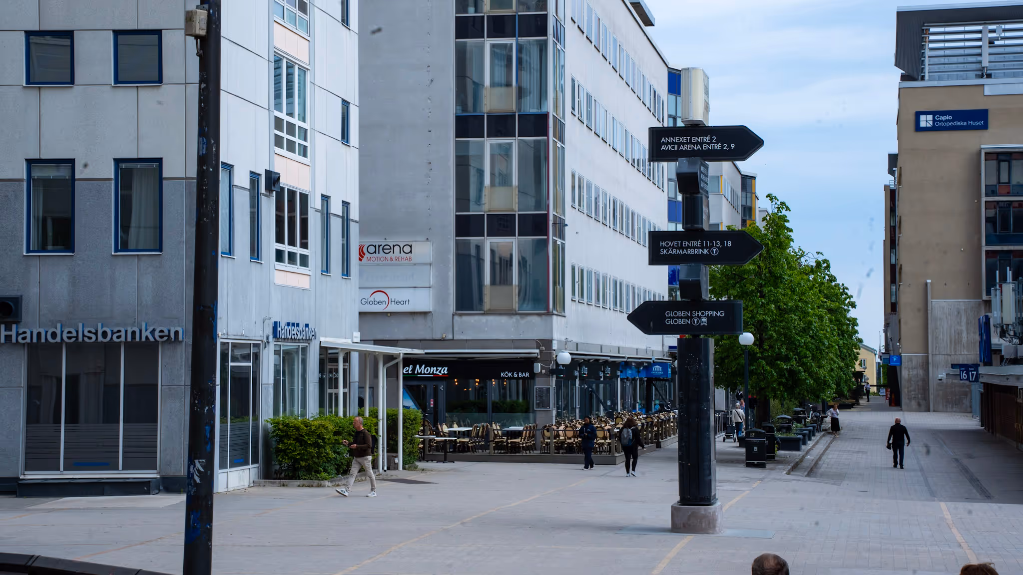 En stadsgata med gående människor, skyltar som pekar mot arenor och restauranger vid modern byggnad.