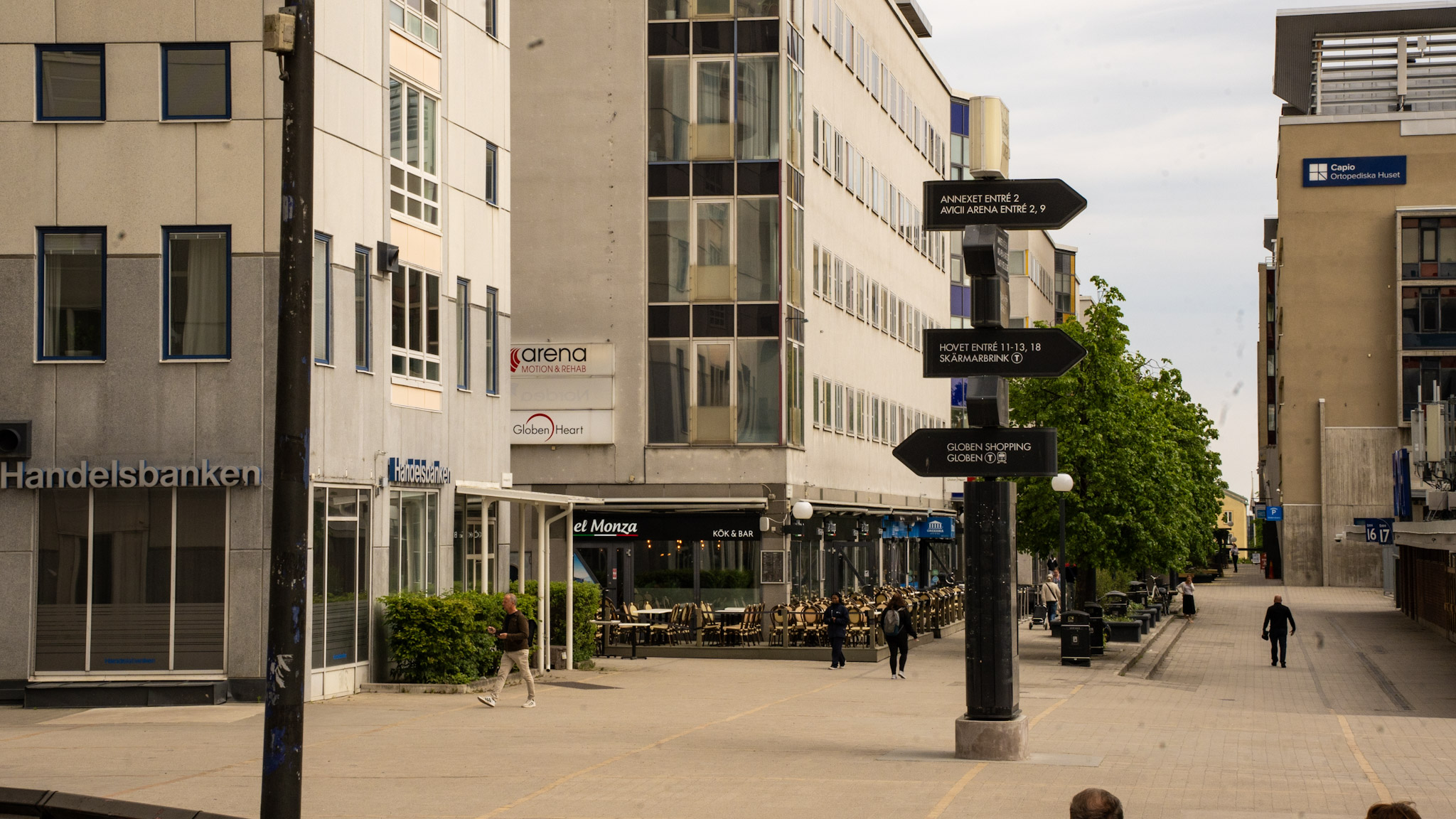 Gågata vid kontorsbyggnader med kafé och skyltar som pekar mot arenan och shopping.