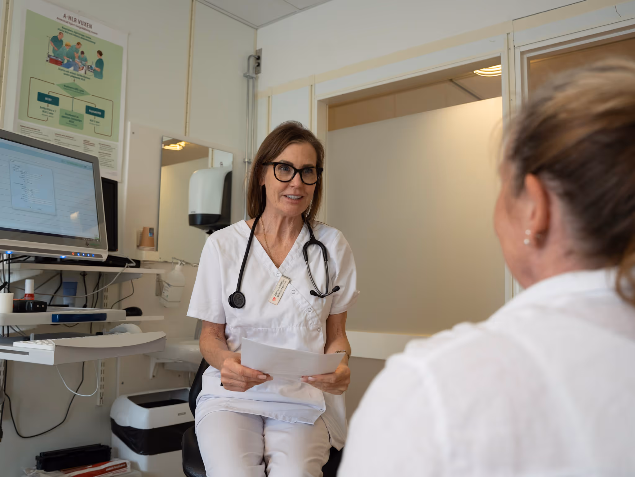Sjuksköterska i vitt samtalar med patient i ett läkarrum med dator och medicinska affischer.