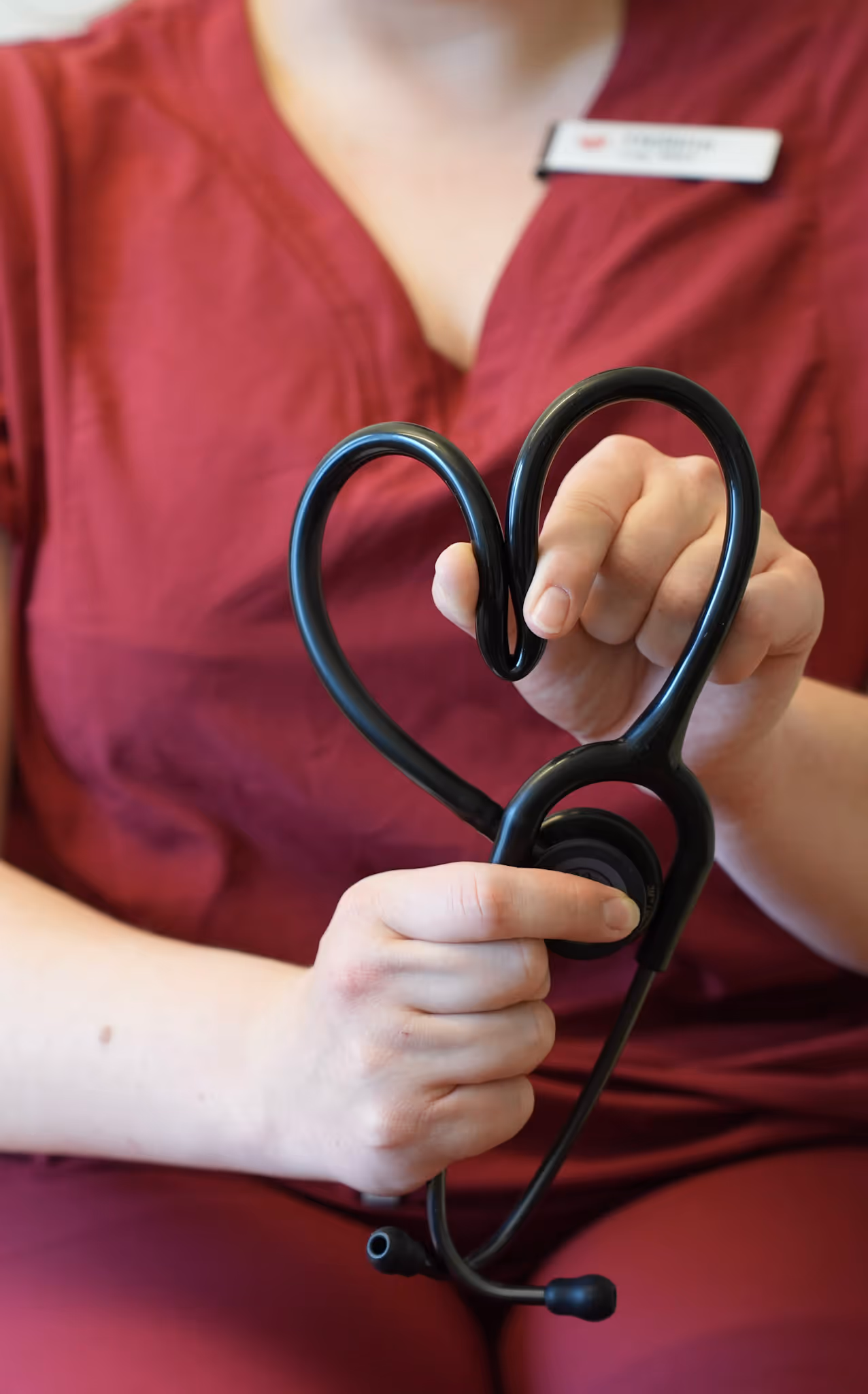 Person in burgundy scrubs holding a black stethoscope shaped like a heart.